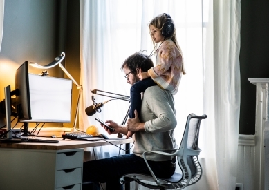 Father sitting at computer desk and working on phone while daughter sits on his shoulders listening to music.